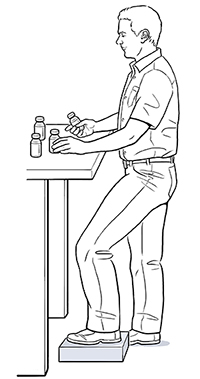 Man standing at work table with one foot elevated on a block.