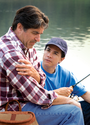Man sitting next to lake with fishing rod, holding arm and looking distressed. Younger man sitting next to him, looking concerned.