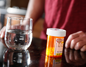 Closeup of pill bottle and glass of water with man in background.
