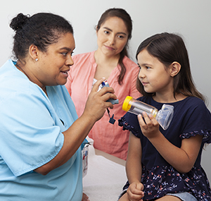 Healthcare provider showing girl how to use metered-dose inhaler with spacer and mask. Woman standing nearby.