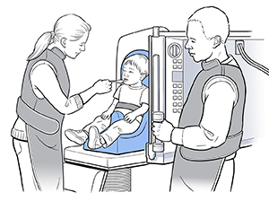 Hay un niño pequeño sentado en un asiento para niños en un aparato de radiografía. Una proveedora de atención médica le da de comer al niño con un cuchara. Otro proveedor de atención médica está cerca de pie.