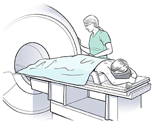 Una mujer acostada boca abajo sobre una camilla de RM. La camilla está lista para entrar en el tubo de RM. Una proveedora de atención médica está de pie junto a la mujer.